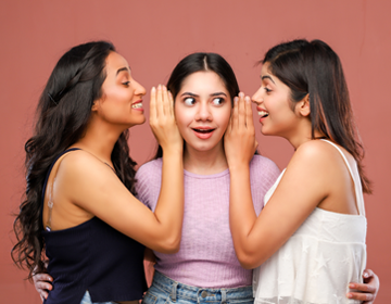 Trzy młode kobiety stojące razem, dwie z nich szepczą do trzeciej, symbol rozmowy i dzielenia się doświadczeniami.